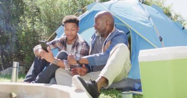 Mutlu Afro-Amerikan baba ve yetişkin oğul dışarıda oturmuş akıllı telefon kullanıyorlar, ağır çekimde. İletişim, kamp, boş zaman, babalık, birliktelik, aile ve açık hava yaşam tarzı, değişmemiş..