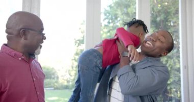 Mutlu Afro-Amerikan baba, oğul ve büyükbabanın portresi güneşli odada kucaklaşıyor, ağır çekimde. Aile, birliktelik, mutluluk, yaşam tarzı ve aile hayatı değişmedi..