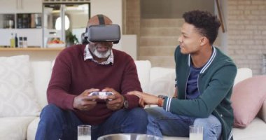 Mutlu Afro-Amerikan babası evde yetişkin oğluyla video kulaklık ve oyun sahası kullanıyor, ağır çekimde. İletişim, video oyunu, boş zaman, teknoloji, eğlence, babalık, aile ve aile hayatı, değiştirilmemiş.