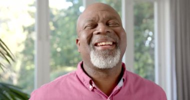 Mutlu son sınıf Afro-Amerikalı bir adamın portresi güneşli oturma odasında gülüyor, ağır çekimde. Yaşlılar, emekliler, mutluluklar ve aile hayatı, değişmemiş..