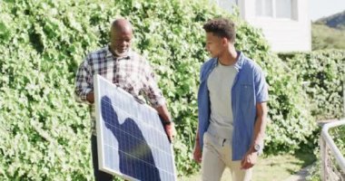 Afrika kökenli Amerikalı baba elinde güneş paneli tutarak güneşli bahçede oğluyla konuşuyor, ağır çekimde. Güneş enerjisi, fotovoltaik panel, öğrenme, birliktelik, babalık, aile ve yaşam tarzı, değişmemiş..