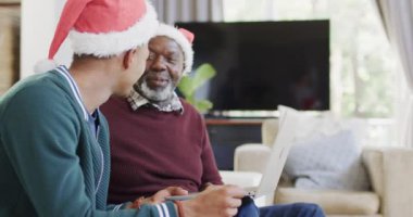 Mutlu Afro-Amerikan baba ve yetişkin oğul noel şapkaları içinde dizüstü bilgisayardan arıyorlar, ağır çekimde. İletişim, babalık, aile, Noel, gelenek, kutlama ve yaşam tarzı, değiştirilmemiş.