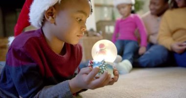 Noel şapkalı Afro-Amerikan oğlu elinde parlayan kar küresi, arka planda aile, ağır çekim. Çocukluk, Noel, kutlama, aile ve gelenek, değiştirilmemiş.