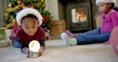 Noel şapkalı Afro-Amerikan oğlu elinde parlayan kar küresi, arka planda aile, ağır çekim. Çocukluk, Noel, kutlama, aile ve gelenek, değiştirilmemiş.