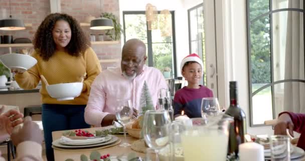 ハッピーアフリカ系アメリカ人の母親は クリスマスのディナーテーブルで多世代の家族に料理を提供しています クリスマス お祝い トータルネスと家庭生活 変わらない — ストック動画
