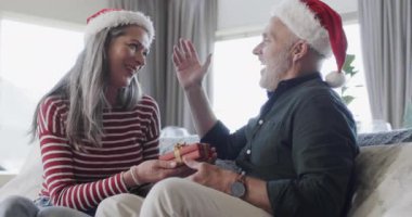 Noel Baba şapkalı orta yaşlı beyaz çift Noel 'de evde hediye veriyorlar, ağır çekimde. Noel, gelenek, kutlama, yaşam tarzı ve ev hayatı, kopya alanı, değişmemiş.