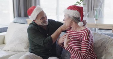 Noel Baba şapkalı orta yaşlı beyaz çift Noel 'de evde sarılıyorlar, ağır çekimde. Noel, gelenek, kutlama, yaşam tarzı ve ev hayatı, kopya alanı, değişmemiş.