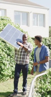 Güneş paneli olan ve oğlu güneşte konuşan Afrikalı Amerikalı bir babanın dikey videosu, yavaş çekim. Güneş enerjisi, fotovoltaik panel, enerji, babalık, aile, ev hayatı ve yaşam tarzı, değiştirilmemiş.