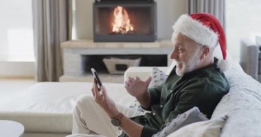 Noel Baba şapkalı orta yaşlı beyaz adam Noel 'de evde akıllı telefon kullanıyor, ağır çekimde. İletişim, Noel, gelenek, kutlama, yaşam tarzı ve ev hayatı, kopyalama alanı, değiştirilmemiş.