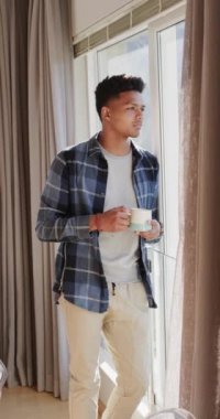 Afro-Amerikan bir adamın güneşli pencereden kahve içerken çekilmiş dikey videosu, yavaş çekim. Düşünce, rahatlama, ev hayatı ve yaşam tarzı, değişmemiş..