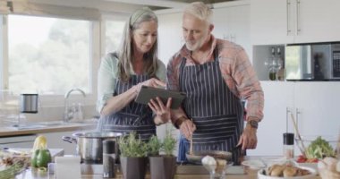 Tabletli orta yaşlı beyaz bir çift, yemek hazırlıyorlar, mutfakta yemek pişiriyorlar, ağır çekimde. İlişki, yemek, yemek, yaşam tarzı ve ev hayatı, kopyalama alanı, değiştirilmemiş.