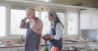 Orta yaşlı beyaz bir çift önlük takıyor, mutfakta yemek pişiriyor, ağır çekimde. İlişki, yemek, yemek, yaşam tarzı ve ev hayatı, kopyalama alanı, değiştirilmemiş.