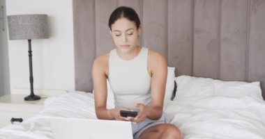 Çift ırklı kadın dizüstü bilgisayar ve akıllı telefon kullanarak yatakta oturuyor, ağır çekimde. Yaşam tarzı, iletişim ve aile hayatı, değişmedi..