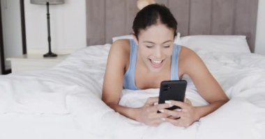Çift ırklı mutlu kadın akıllı telefon kullanarak yatağa uzanıyor, ağır çekimde. Yaşam tarzı, iletişim ve aile hayatı, değişmedi..