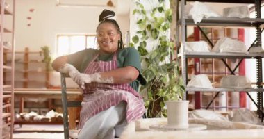 Çömlek atölyesinde oturan ve gülümseyen Afro-Amerikan kadın çömlekçi. Çömlek, seramik, el yapımı, yerel işletme, hobiler ve el işi, değiştirilmemiş.
