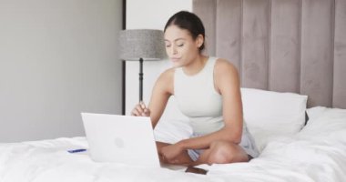 Çift ırklı bir kadın dizüstü bilgisayar kullanarak yatakta oturuyor, ağır çekimde. Yaşam tarzı, iletişim ve aile hayatı, değişmedi..