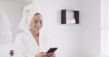 Kafasında havluyla banyoda akıllı telefon kullanan mutlu çift ırklı kadın portresi, yavaş çekim. Yaşam tarzı, kişisel bakım, iletişim ve ev hayatı, değişmemiş..