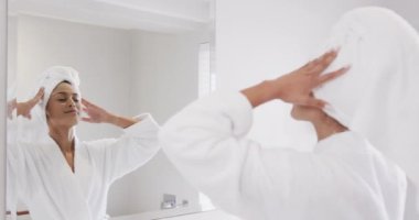 Kafasına havlu geçirmiş mutlu bir çift ırklı kadın banyoda aynaya bakıyor, ağır çekimde. Yaşam tarzı, kişisel bakım ve ev hayatı, değişmemiş..