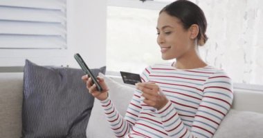 Kanepede oturan mutlu melez kadın akıllı telefonu online alışveriş için kullanıyor, ağır çekimde. Yaşam tarzı, iletişim, alışveriş ve ev hayatı, değişmedi..