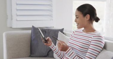 Kanepede oturan mutlu melez kadın akıllı telefonu online alışveriş için kullanıyor, ağır çekimde. Yaşam tarzı, iletişim, alışveriş ve ev hayatı, değişmedi..