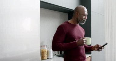 Kahve içen ve mutfakta akıllı telefon kullanan mutlu çift ırklı adam, yavaş çekim. Ev hayatı, teknoloji ve yaşam tarzı, değişmedi..