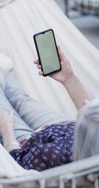 Kafkasyalı yaşlı kadının hamakta fotokopi alanı olan akıllı telefonu yavaş çekimde kullandığının dikey videosu. Kıdemli yaşam tarzı, iletişim, yaz ve tatil, değişmeden.