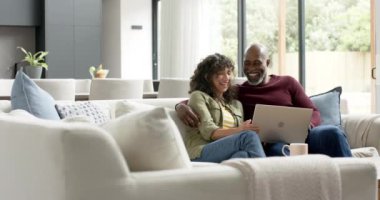 Çift ırklı mutlu çift koltukta oturup dizüstü bilgisayar kullanıyor, ağır çekimde. Ev hayatı, teknoloji ve yaşam tarzı, değişmedi..