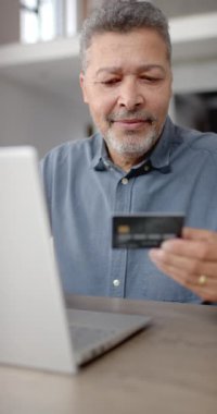Üst düzey melez bir adamın dizüstü bilgisayar kullanırken ve evde dikey kredi kartı kullanırken çekilmiş dikey videosu. Emeklilik, eğlence, teknoloji, iletişim, alışveriş ve ev hayatı kavramı.