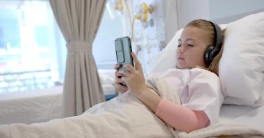 Kafkasyalı hasta hastane yatağında kulaklıklarıyla akıllı telefon, fotokopi odası ve ağır çekim kullanıyor. İletişim, teknoloji, çocukluk, hastalık, sağlık hizmetleri, sağlık ve hastane, değiştirilmemiş.