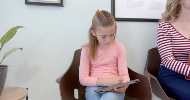 病院の受付で母親と座っているタブレットを使用している白人少女の患者 ゆっくりとした動き コミュニケーション 幼少期 医療サービス 変わらない — ストック動画