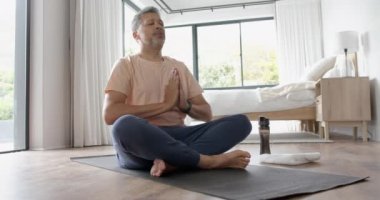 Evde yoga meditasyonu yapan yaşlı, çift ırklı rahat bir adam, ağır çekimde. Lotus pozisyonu, emeklilik, yoga, refah, egzersiz ve sağlıklı yaşam tarzı, değişmemiş.