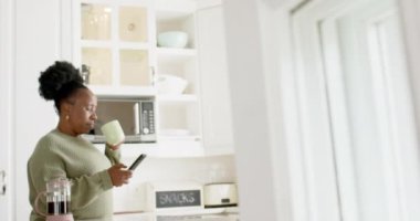 Mutlu Afro-Amerikan son sınıf öğrencisi kadın kahve içiyor, güneşli mutfakta akıllı telefon kullanıyor, ağır çekimde. Ev, emeklilik ve yaşlı yaşam tarzı, ev hayatı, rahatlama, iletişim, değişmemiş.