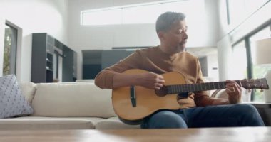 Odaklanmış yaşlı çift ırklı adam koltukta oturup gitar çalıyor, boşluğu kopyalıyor, yavaş çekim yapıyor. Müzik, rahatlama, yaratıcılık, hobiler, emeklilik, ev hayatı ve yaşlılar yaşam tarzı, değiştirilmemiş.