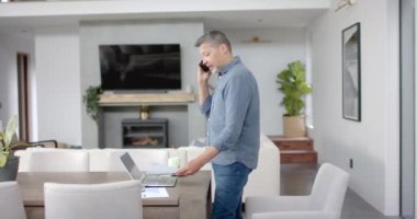 Telefonda kıdemli melez adam, evrak işlerine bakıyor, ev işi, kopyalama alanı, ağır çekim. Uzaktan çalışma, iş, iletişim, ev hayatı ve yaşlı yaşam tarzı, değiştirilmemiş.