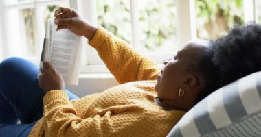 Mutlu Afro-Amerikan son sınıf öğrencisi kadın yalan söylüyor, kitap okuyor ve güneşli odada gülümsüyor, ağır çekimde. Ev, emeklilik ve yaşlı yaşam tarzı, boş zaman ve ev hayatı, rahatlama, refah, değişmemiş.