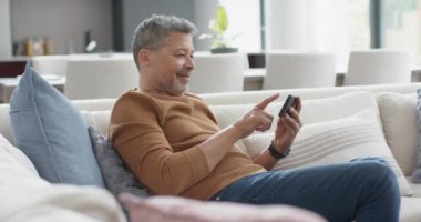Güneşli oturma odasında akıllı telefon kullanan kıdemli çift ırklı mutlu bir adam. Ağır çekimde. İletişim, emeklilik, ev hayatı ve yaşlı yaşam tarzı, değiştirilmemiş.