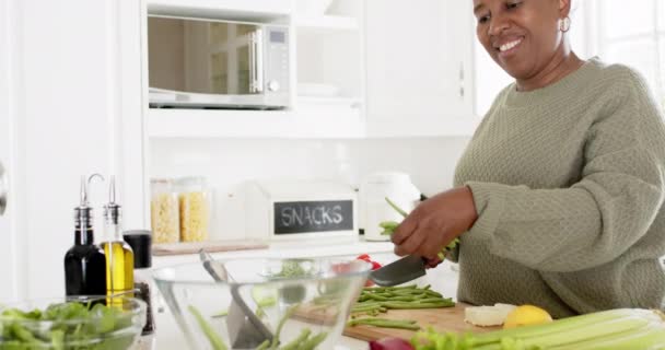 ハッピーアフリカ系アメリカ人のシニア女性が野菜をチョッピングし 晴れたキッチンで微笑み ゆっくりとした動き ホーム 退職とシニアライフスタイル 家庭生活 食べ物 変更されていない — ストック動画