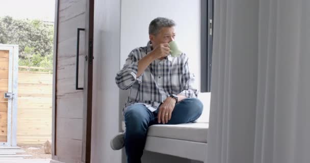 Avslappnad Senior Biracial Man Sitter Vid Fönstret Och Dricker Kaffe — Stockvideo