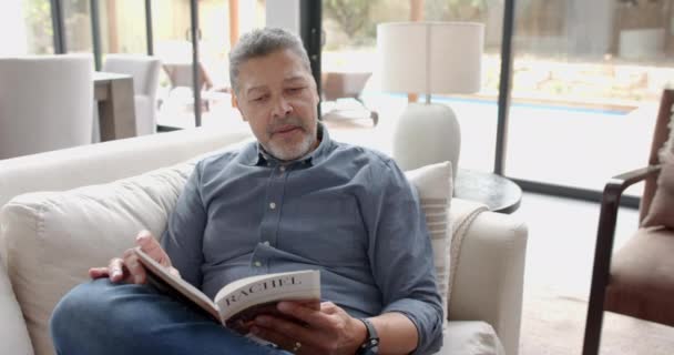 Сосредоточен Старшем Расовом Человеке Сидящем Диване Читающем Книгу Гостиной Дома — стоковое видео