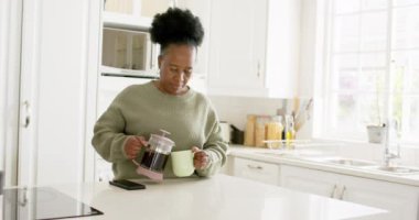 Mutlu Afro-Amerikan son sınıf öğrencisi kadın kahveyi koyuyor ve güneşli mutfakta gülümsüyor, ağır çekimde. Ev, emeklilik ve yaşlı yaşam tarzı, boş zaman ve ev hayatı, rahatlama, refah, değişmemiş.
