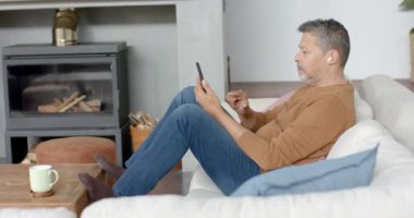 Oturma odasındaki koltukta oturan akıllı telefon ve kulaklık kullanan kıdemli bir melez. İletişim, rahatlama, emeklilik, ev hayatı ve yaşlılar yaşam tarzı, değişmedi..