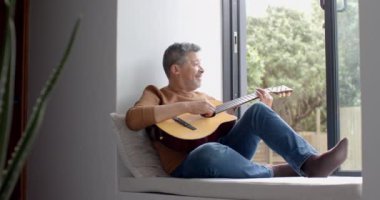 Açık pencerede oturan, gitar çalan, mekanı kopyalayan, ağır çekim mutlu kıdemli çift ırklı adam. Müzik, rahatlama, yaratıcılık, hobiler, emeklilik, ev hayatı ve yaşlılar yaşam tarzı, değiştirilmemiş.