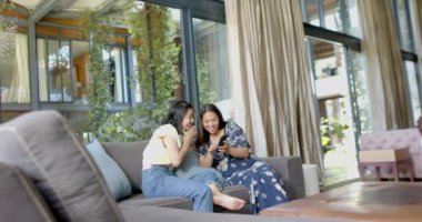 Mutlu Asyalı bayan arkadaşlar akıllı telefon kullanıyor ve güneşli oturma odasında gülümsüyor, ağır çekimde. Yaşam tarzı, boş zaman, refah ve ev hayatı, arkadaşlık, iletişim, değişmemiş.