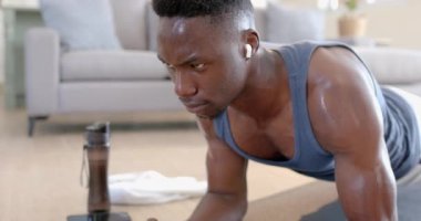 Yorgun Afro-Amerikan bir adam güneşli oturma odasında kalas yapıyor ve kulaklık takıyor, ağır çekimde. Ev, egzersiz, fitness ve sağlıklı yaşam tarzı, iletişim, teknoloji, değişmemiş.