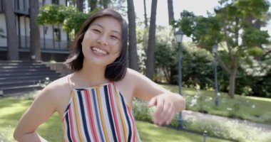 Siyah düz saçlı mutlu Asyalı kadın güneşli bahçede ayakta ve gülümsüyor, ağır çekimde. Yaz, tatil, doğa, refah, değişmemiş.