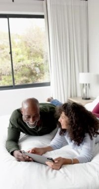 Tablet, kopyalama alanı ve ağır çekim kullanarak yatağa uzanan mutlu çiftin dikey videosu. İletişim, rahatlama, ilişki, birliktelik, ev hayatı ve yaşam tarzı, değişmemiş..