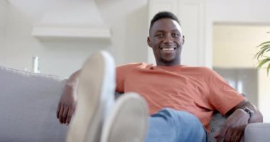 Mutlu Afro-Amerikalı adam güneşli oturma odasında oturuyor ve gülümsüyor, ağır çekimde. Boş zaman ve ev hayatı, duygu, mutluluk, refah, değişmemiş.