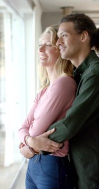 Evdeki pencerede birbirini kucaklayan mutlu çiftin dikey videosu, yavaş çekim. Ev hayatı ve yaşam tarzı değişmedi..