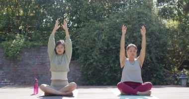 Odaklanmış Asyalı bayan arkadaşlar güneşli terasta yoga meditasyonu yapıyorlar, yavaş çekim. Sağlıklı yaşam tarzı, zindelik ve refah, arkadaşlık, değişmemiş.