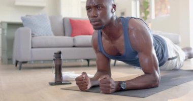 Yorgun Afro-Amerikan bir adam oturma odasında kalas, kulaklık ve akıllı telefon kullanıyor, ağır çekimde. Ev, egzersiz, fitness ve sağlıklı yaşam tarzı, iletişim, teknoloji, değişmemiş.